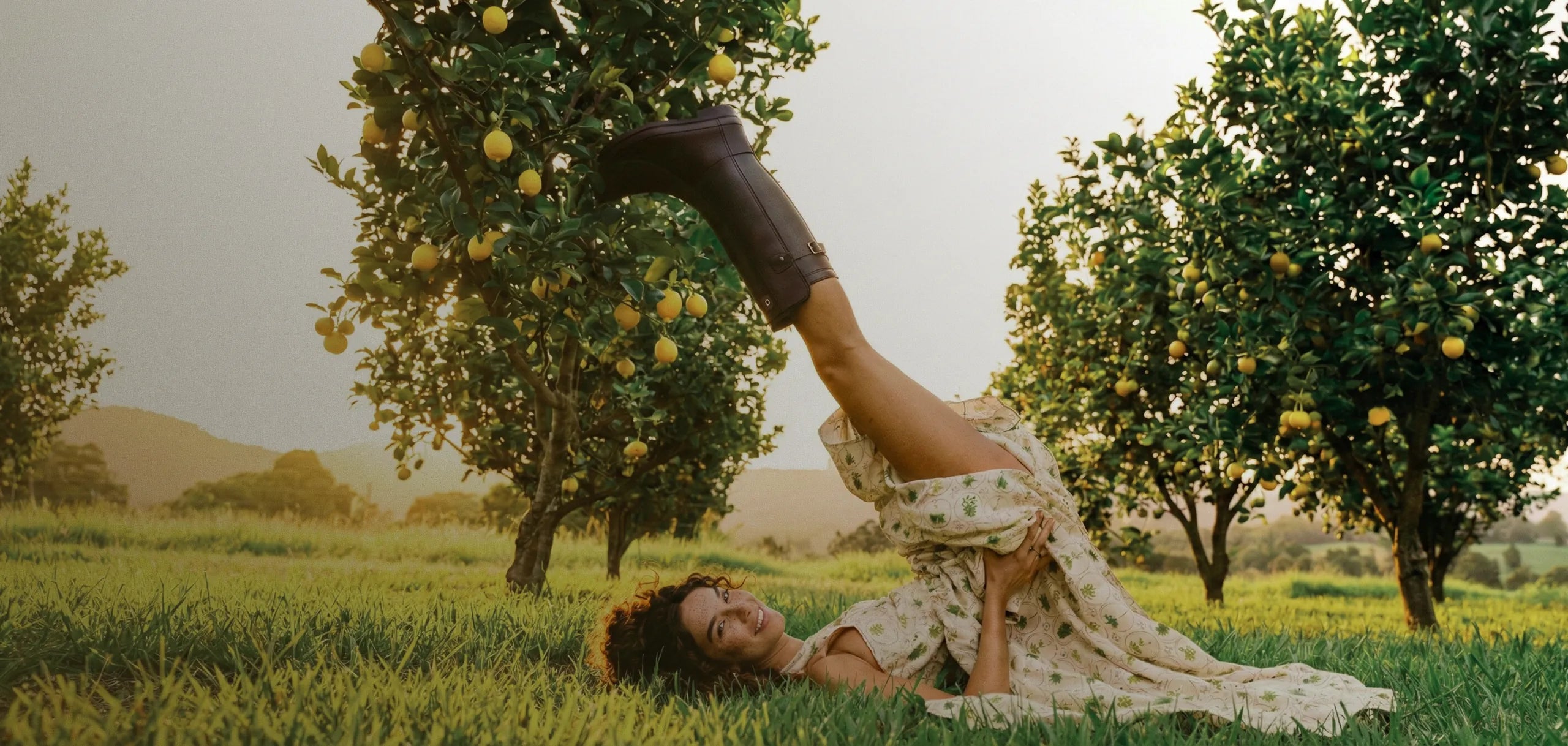 Woman in a dress and boots lying on grass with lemon trees in the background
